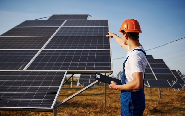 Mostrando de la mano. Trabajador de sexo masculino en uniforme azul al aire libre con baterías solares en un día soleado.