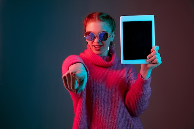 Mostrando la pantalla de la tableta en blanco. Retrato de mujer caucásica aislado sobre fondo de estudio degradado en luz de neón. Modelo de mujer hermosa con el pelo rojo. Concepto de emociones humanas, expresión facial, anuncio.