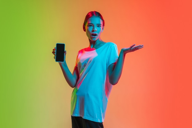 Mostrando smartphone. Retrato de jovem caucasiana em fundo gradiente estúdio verde-laranja em luz de néon. Conceito de juventude, emoções humanas, expressão facial, vendas, anúncio. Linda modelo adolescente.