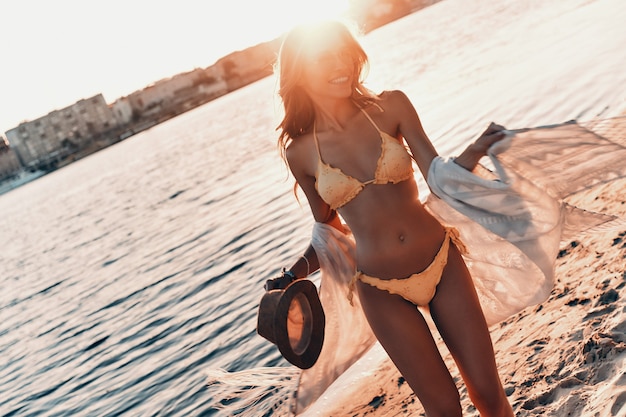 Mostrando su increíble cuerpo. Atractiva mujer joven sonriendo mientras está de pie en la playa al aire libre