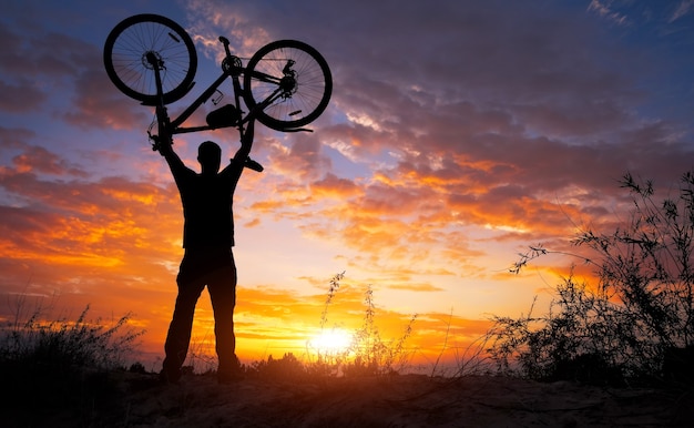 Mostre a silhueta do homem em ação, levantando uma bicicleta acima da cabeça na campina com o pôr do sol