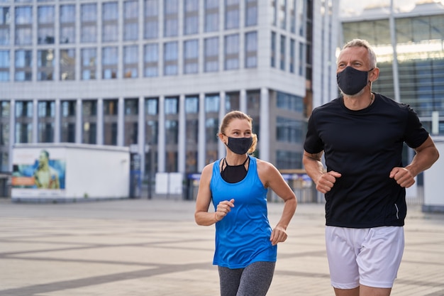 Motivado pareja de mediana edad mujer y hombre con máscaras protectoras trotar al aire libre en la ciudad para