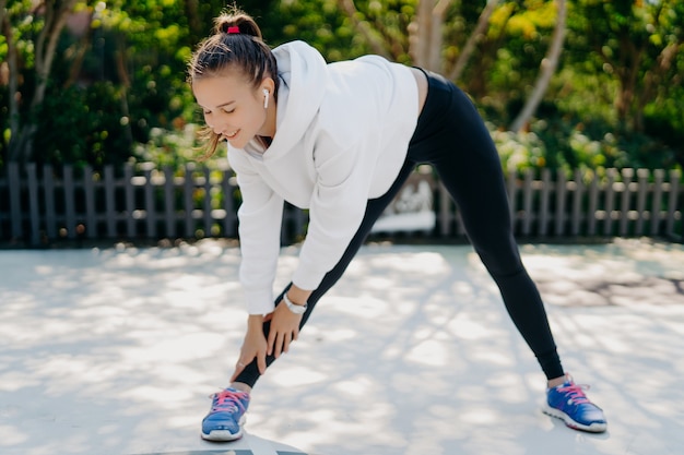 Motivierte junge erwachsene Frau treibt regelmäßig Sport reicht Hand zu Fuß beugt sich nach unten hat Füße schulterbreit auseinander trägt Sportkleidung trainiert gerne Outdoor-Sportaktivitäten zur Gewichtsreduktion