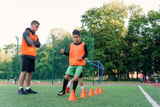 Motivierte Jungen in orangefarbenen Westen, die während des Fußballtrainings im Stadion zwischen den Plastikkegeln laufen.