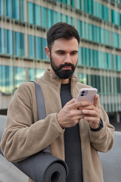 Motivierter sportlicher Mann mit dickem Bart konzentriert auf Smartphone-Bildschirm liest Artikel online trägt Fitnessmatte und trainiert im Freien Posen gegen moderne Gebäudekontrollen Newsfeed