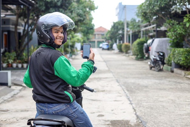 Moto taxista asiática en línea con chaqueta verde y casco sonriendo mientras sostiene el teléfono inteligente