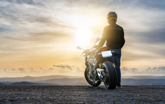 Motociclista ao lado da motocicleta, observando o pôr do sol - copie o espaço.