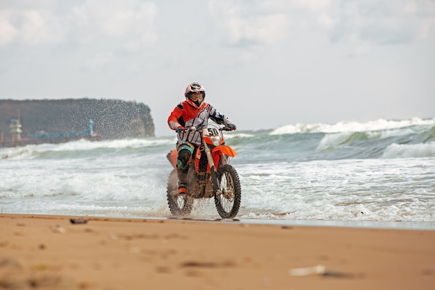 Motociclista em um traje de proteção anda de moto no mar, salpicos voam debaixo das rodas.