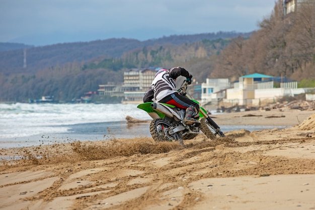 Motociclista em um traje de proteção anda de moto no mar, salpicos voam debaixo das rodas.