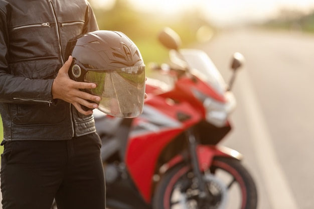 Motociclista guapo llevar chaqueta de cuero, sosteniendo el casco en la carretera