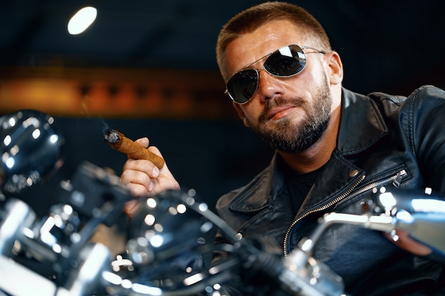 Motociclista legal homem barbudo em óculos de sol, sentado em sua moto e fumando charuto
