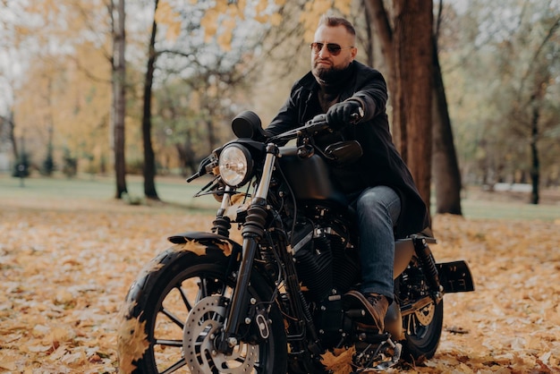 Motociclista masculino dirige na natureza em bicicleta rápida usa óculos escuros jaqueta preta luvas jeans e botas aproveita a temporada de outono passa o tempo livre ativamente pronto para uma longa viagem transporte de pessoas dirigindo