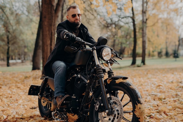Motociclista masculino dirige na natureza em bicicleta rápida usa óculos escuros jaqueta preta luvas jeans e botas aproveita a temporada de outono passa o tempo livre ativamente pronto para uma longa viagem transporte de pessoas dirigindo