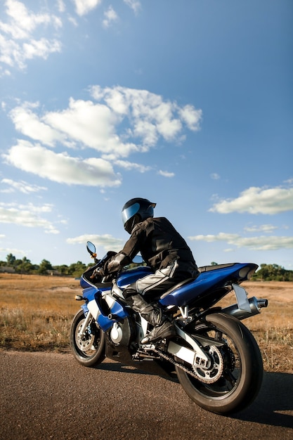 Motociclista ou motociclista em um terno de couro preto e capacete senta-se em uma motocicleta esportiva e olha para a distância.