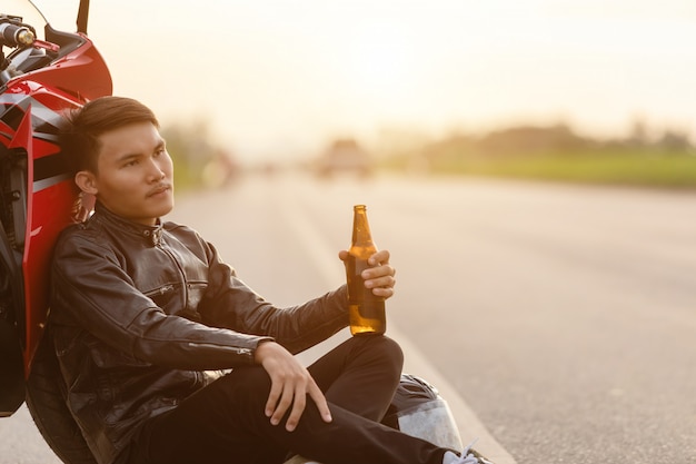 Motociclista sentado na estrada ao lado de sua moto e bebendo cerveja