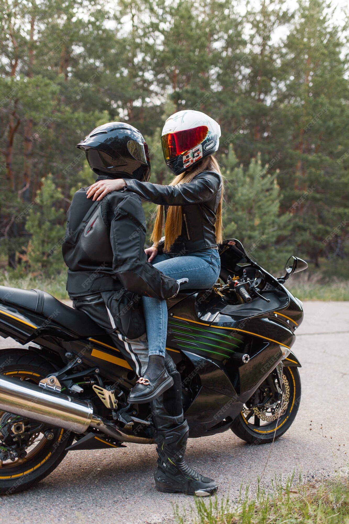 Motociclista en traje de cuero se con una chica en una motocicleta en cascos y abrazos contra el fondo de árboles en el bosque | Foto Premium