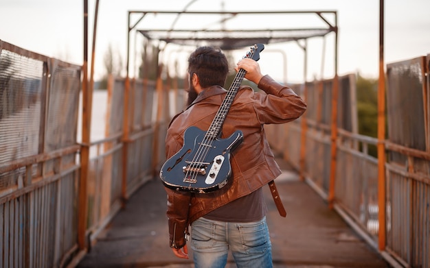 Motoqueiro barbudo brutal com cabelo grisalho, uma jaqueta de couro marrom e jeans azul está de costas e segura uma guitarra elétrica preta no ombro