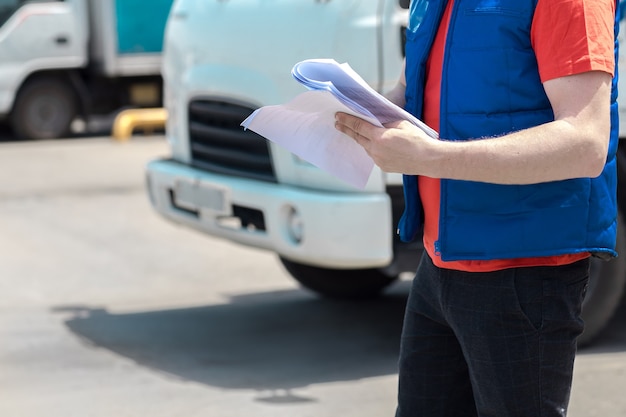 Motorista de correio fardado fazendo anotações em documento e caminhão branco de entrega atrás dele.