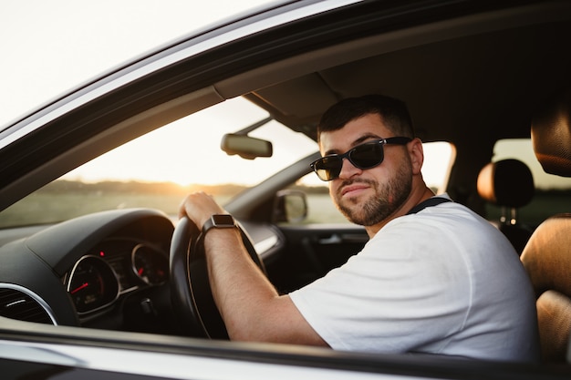 Motorista de homem barbudo usando óculos escuros, sentado no carro e olhando para a câmera