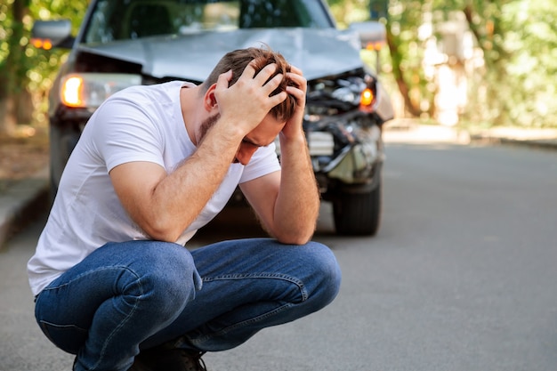 Motorista de jovem branco em acidente de carro e segurando sua cabeça perto de um carro quebrado na estrada após um acidente de carro. homem caucasiano, rosto, palma da mão segurando ferimento na cabeça após acidente