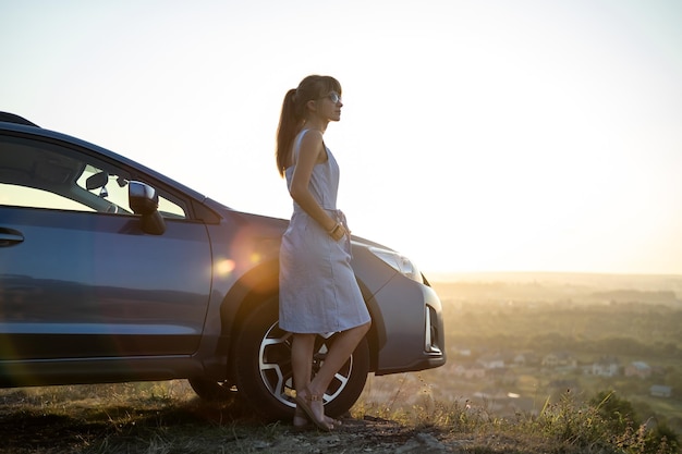 Motorista de mulher feliz em vestido azul de verão aproveitando a noite quente perto de seu carro Conceito de viagens e férias