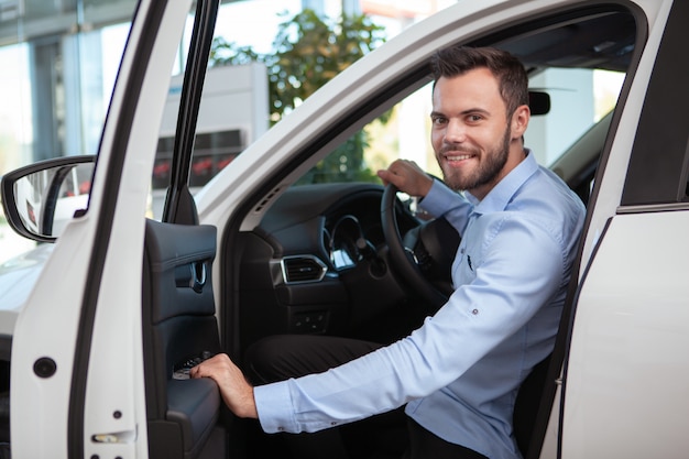 Motorista do sexo masculino bonito alegre sorrindo para a câmera, sentado em um carro novo na concessionária