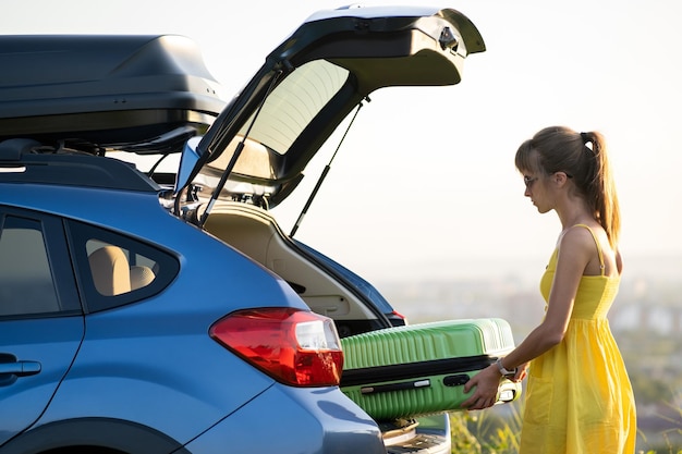 Motorista feminina em vestido de verão colocando mala verde dentro do porta-malas do carro Conceito de viagens e férias