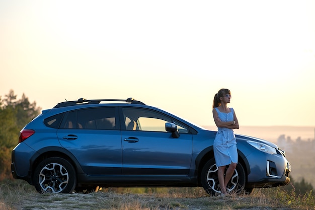 Foto motorista jovem feliz em vestido azul, aproveitando a noite quente de verão em pé ao lado de seu carro. conceito de viagens e férias.