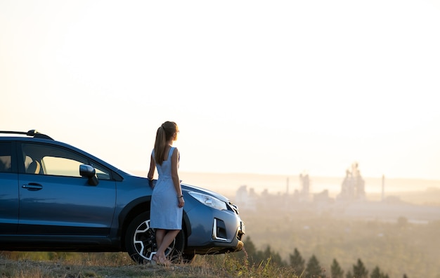 Motorista jovem feliz em vestido azul encostado em seu carro, aproveitando o dia quente de verão.