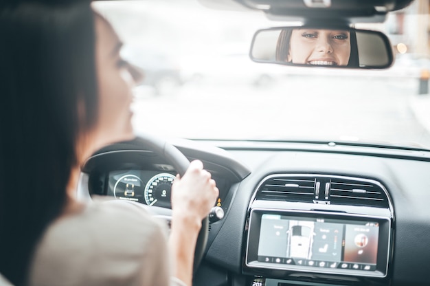 Motorista no espelho retrovisor. Retrovisor de uma jovem atraente em roupa casual olhando no espelho retrovisor e sorrindo enquanto dirige um carro