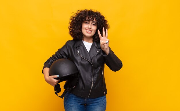 Motorradfahrerfrau lächelt und sieht freundlich aus, zeigt Nummer drei oder dritte mit der Hand nach vorne, Countdown