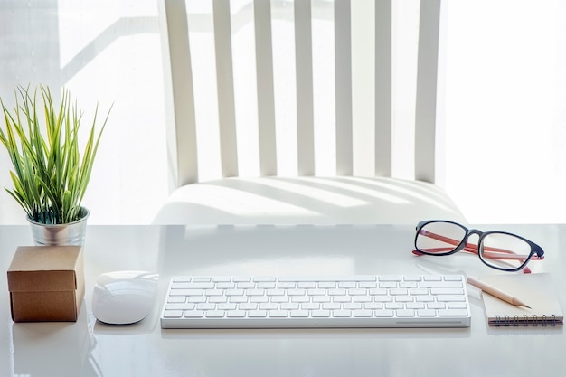 mouse, nota en blanco, lápiz, teclado blanco, anteojos y houseplant en mesa blanca