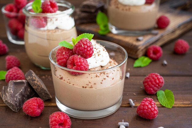 Mousse de chocolate con crema batida y frambuesas frescas en un vaso sobre un fondo de madera oscura.