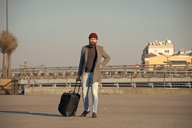 Movendo-se para a nova cidade sozinho Hipster pronto para desfrutar de viagem Carregar bolsa de viagem Homem barbudo viagem hipster com bolsa de bagagem nas rodas Viajante com mala chegar ao fundo urbano da estação ferroviária do aeroporto