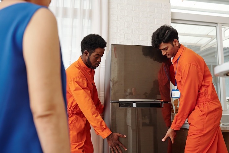 Foto movers em uniforme laranja carregando uma geladeira grande nova para o apartamento de uma senhora idosa