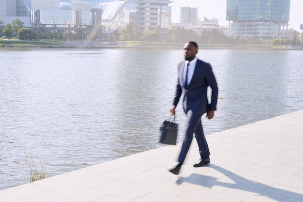 Movimiento borroso del joven empresario africano contemporáneo en traje azul oscuro moviéndose a lo largo de la orilla del río mientras se apresuraba a trabajar por la mañana