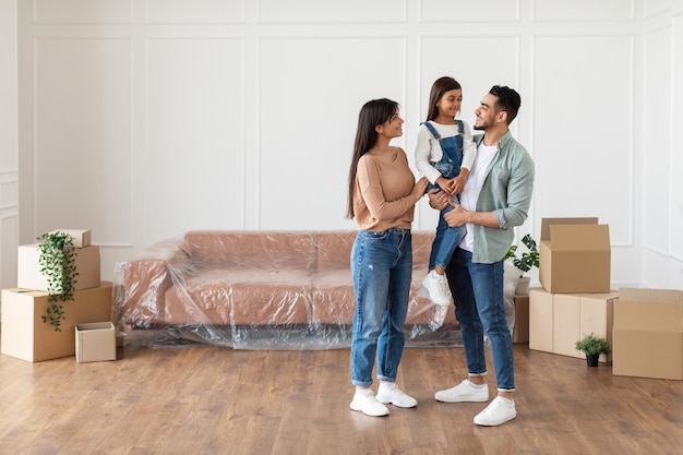 Movimiento de casa, concepto de reparación. Longitud de cuerpo entero de la alegre familia joven de tres personas de pie en el nuevo apartamento vacío, mirando el uno al otro, espacio de copia libre. Padres, tenencia, hija, en, manos