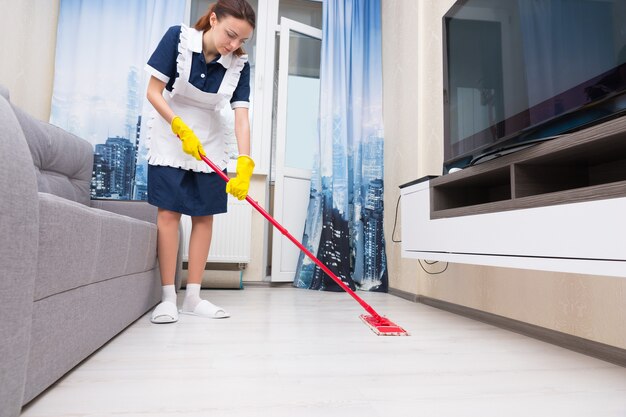 Mucama o ama de llaves en un pulcro delantal blanco limpiando el piso de una sala de estar con un trapeador rojo colorido, vista de ángulo bajo al nivel del piso