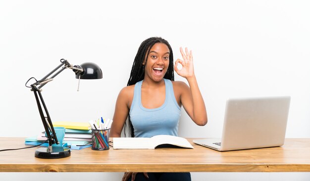 Muchacha adolescente afroamericana del estudiante con el pelo trenzado largo en su lugar de trabajo sorprendida y que muestra la muestra aceptable