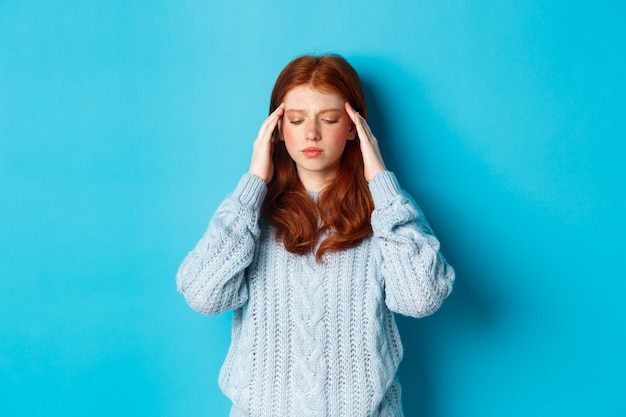 La muchacha adolescente angustiada del pelirrojo que toca la cabeza, mirando hacia abajo con la expresión de la cara preocupada, de pie contra el fondo azul, tiene un problema.