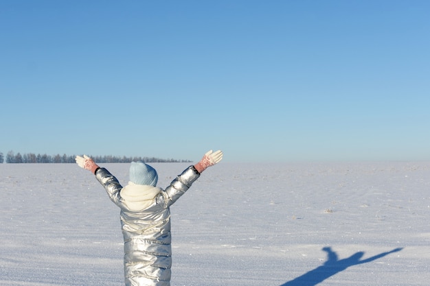 La muchacha adolescente disfruta del invierno, de pie en el campo de nieve levantó las manos al cielo azul.