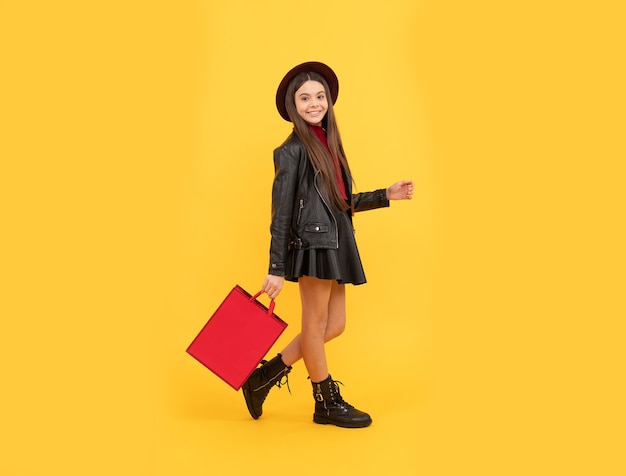 La muchacha adolescente feliz en ropa de cuero y sombrero sostiene el bolso de compras, moda.