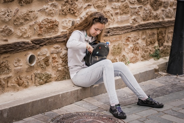 Muchacha adolescente joven que hurga en una mochila que se sienta en los pasos de la casa vieja