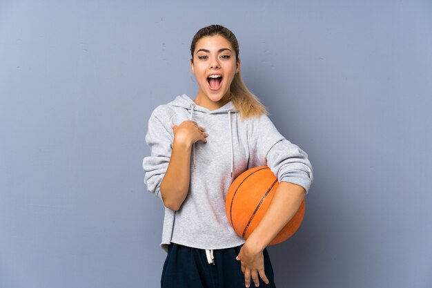 Muchacha del adolescente que juega a baloncesto sobre la pared gris con la expresión facial de la sorpresa
