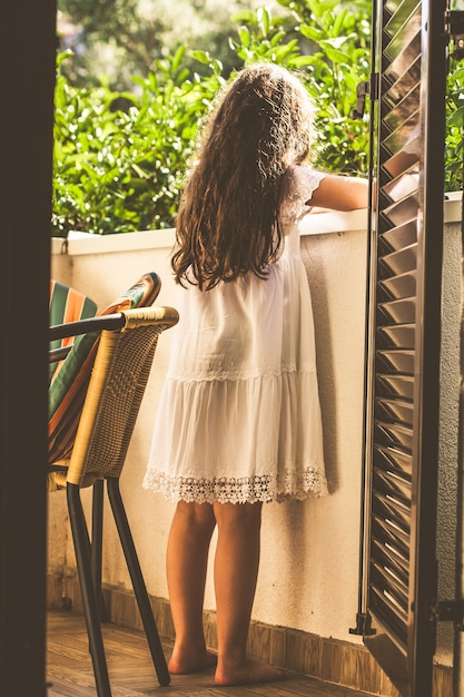 Muchacha adolescente en el vestido blanco y con el pelo marrón largo que se coloca en un balcón en el fondo del jardín