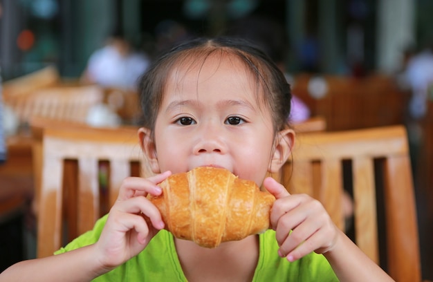 Muchacha adorable del niño asiático que saca una mordedura del cruasán por la mañana en el restaurante.