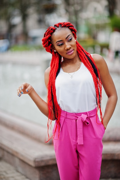 La muchacha afroamericana de moda en pantalones rosados y temores rojos plantea al aire libre contra las fuentes.