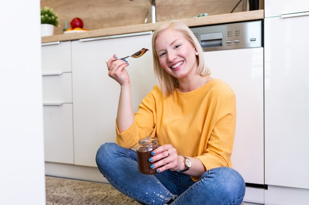 La muchacha albino joven hermosa en ropa casual que prueba el chocolate delicioso se separó mientras que se sentaba en el piso de madera en la cocina doméstica.