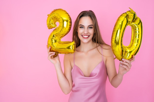 La muchacha alegre del adolescente joven sostiene el globo de la hoja de oro en concepto de la fiesta de cumpleaños del fondo rosado