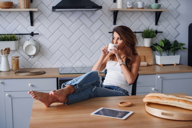 Muchacha alegre en la camiseta blanca usando la tableta mientras que se relaja en una silla en casa en cocina acogedora.